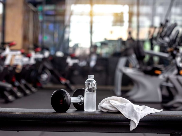 A barbell, water bottle, and white towel are placed on a gym bench, with the martial arts gym out of focus in the background.
