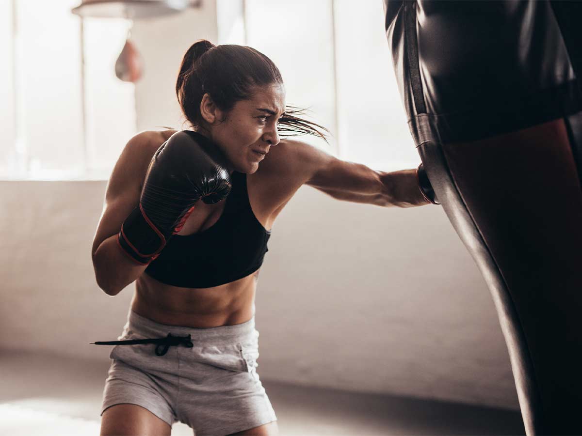 A muscular woman training kickboxing strikes a punching bag full force.