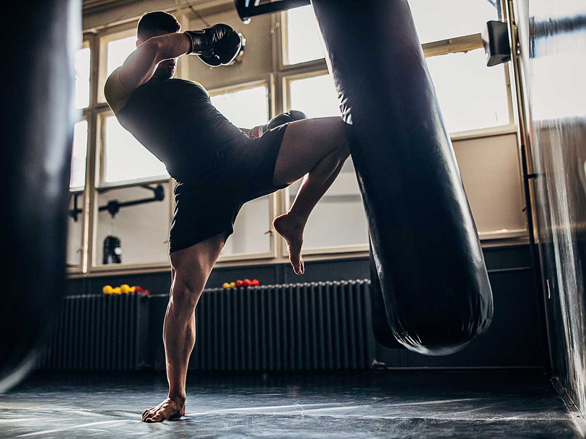 A muscular man performs a high kick against a punching bag while training martial arts.