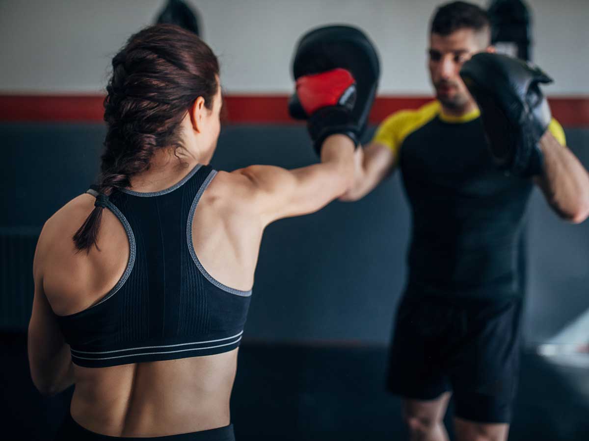 A muscular woman is seen from behind, sparring with a male instructor.