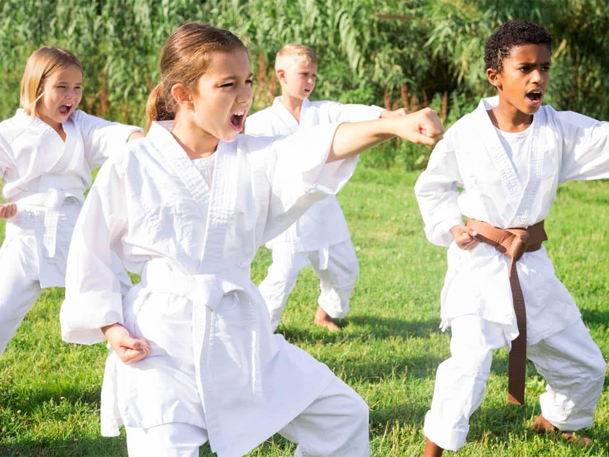 Children in white robes perform taekwondo actions outdoors.